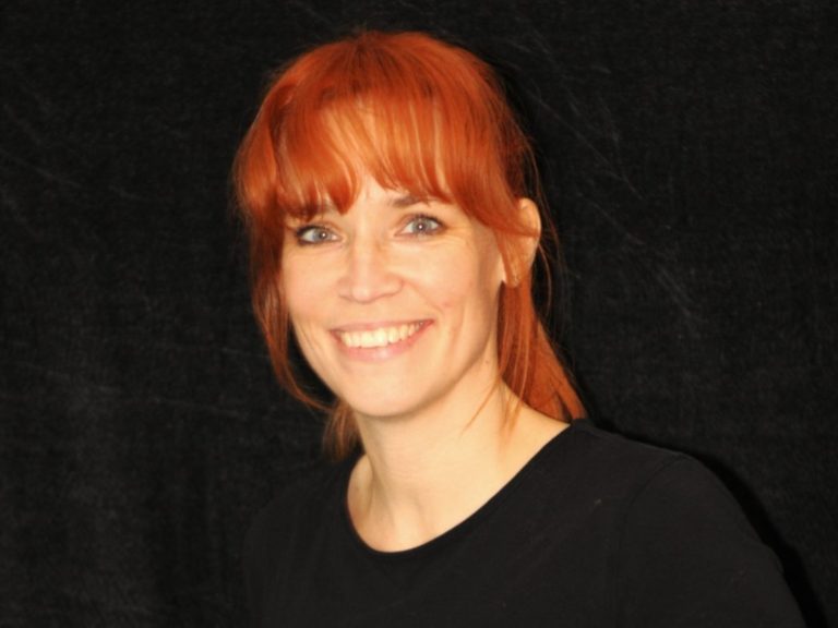 Frau mit rotem Haar und lächelndem Gesicht auf schwarzem Hintergrund.
