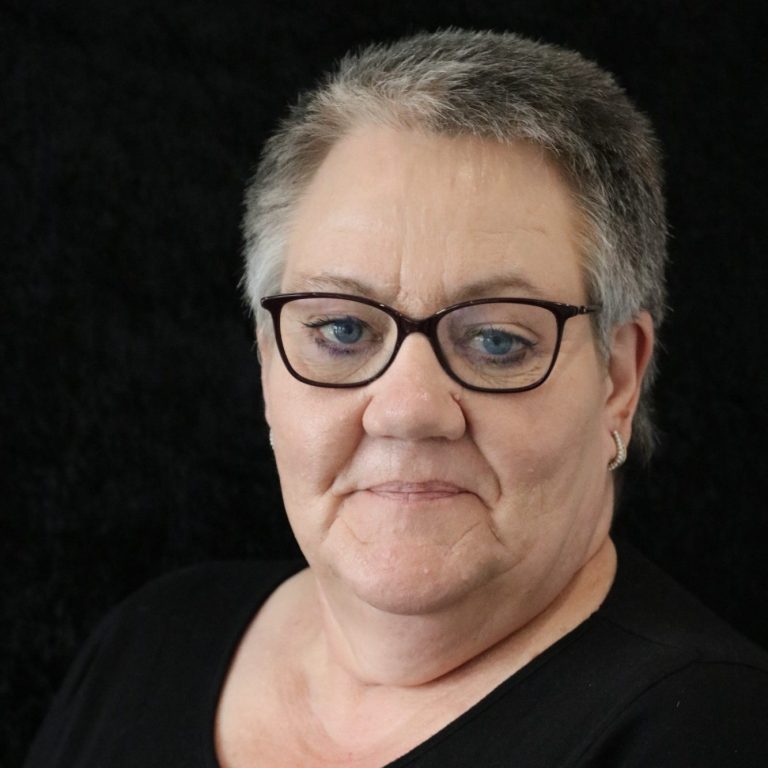 Porträt einer Frau mit Brille, kurzen grauen Haaren und neutralem Gesichtsausdruck.