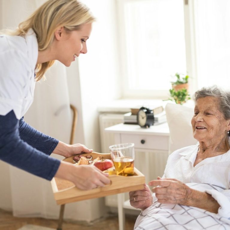 Eine Pflegerin überreicht einer älteren Dame ein Tablett mit Essen und Getränken.
