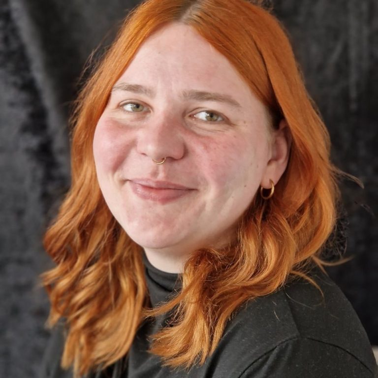 Lächelnde Person mit roten Haaren und schwarzem Oberteil vor schwarzem Hintergrund.