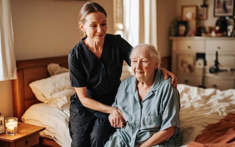 Pflegekraft hilft älterer Frau in einem gemütlichen Schlafzimmer.