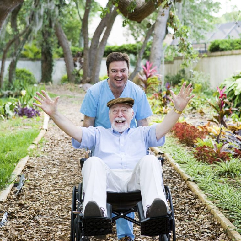 Ein älterer Mann im Rollstuhl wird von einem lächelnden Pfleger in einem Garten geschoben.
