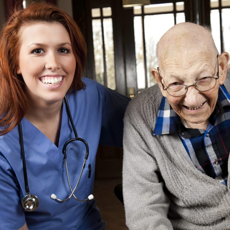 Lächelnde Krankenschwester neben einem älteren Mann bei ihm daheim.